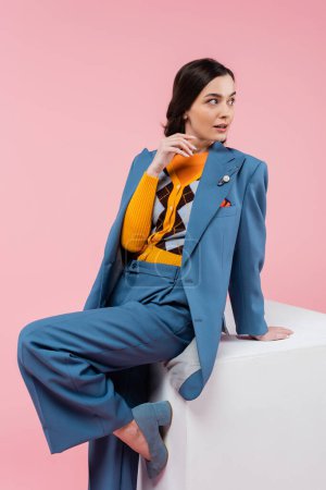 Foto de Bonita mujer en traje pantalón azul posando cerca del cubo blanco y mirando hacia otro lado aislado en rosa - Imagen libre de derechos