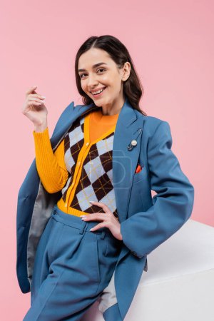 smiling woman in blue suit and bright cardigan posing with hand on hip near white cube isolated on pink