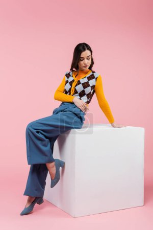 full length of brunette woman in blue pants and trendy cardigan sitting on white cube on pink background