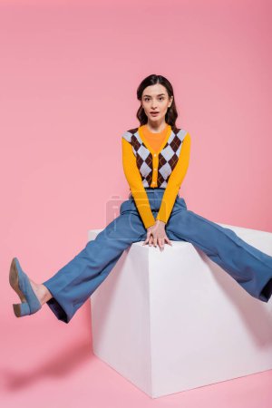 full length of young brunette woman in fashionable outfit looking at camera while sitting on white cube on pink background