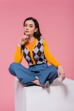 young brunette woman in bright cardigan and blue pants sitting with crossed legs on white cube isolated on pink