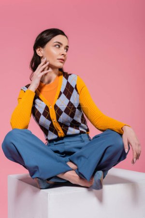 full length of woman in blue pants and orange cardigan with argyle pattern sitting with crossed legs on white cube isolated on pink