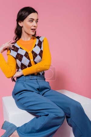 surprised woman in trendy cardigan and blue trousers sitting on white cube and looking away on pink background