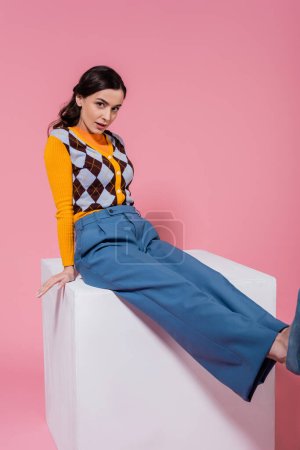 brunette woman in bright cardigan and blue pants sitting with outstretched legs on white cube on pink background