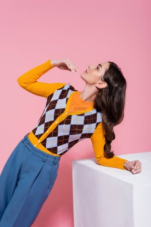 fashionable woman in bright cardigan and blue pants looking up while posing near white cube on pink background