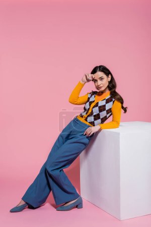 Foto de Longitud completa de la mujer bonita en el cárdigan de moda y pantalones azules apoyados en el cubo blanco sobre fondo rosa - Imagen libre de derechos