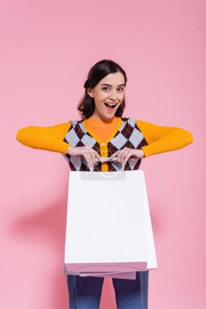 excited woman in orange cardigan with argyle pattern holding white shopping bags and looking at camera on pink background