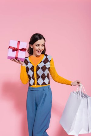 Foto de Mujer alegre en cárdigan de moda y pantalones azules sosteniendo bolsas de compras y caja de regalo mientras mira hacia otro lado sobre fondo rosa - Imagen libre de derechos