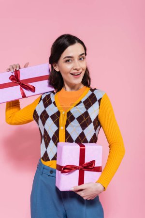 Foto de Mujer morena feliz en cárdigan de moda posando con regalos sobre fondo rosa - Imagen libre de derechos