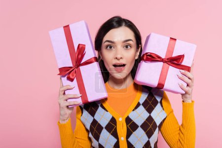 impressed woman in orange cardigan with argyle pattern showing gift boxes while looking at camera isolated on pink