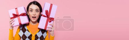 Überraschte Frau in trendiger Strickjacke mit Geschenken und Blick in die Kamera isoliert auf rosa Banner