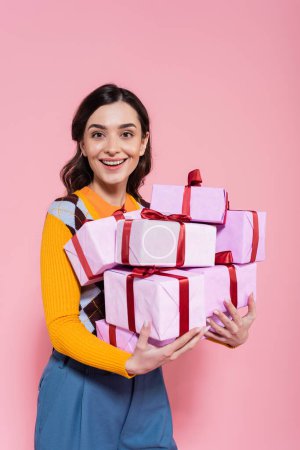 freudige brünette Frau lächelt in die Kamera, während sie Stapel von Geschenken isoliert auf rosa