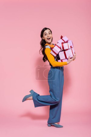 full length of amazed and happy woman in blue pants holding pile of gifts on pink background