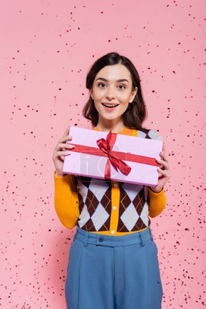 glückliche und stilvolle Frau hält Geschenkbox mit rotem Band auf rosa Hintergrund