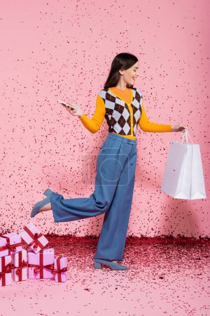 happy woman in trendy clothes posing with shopping bags and mobile phone near gift boxes and confetti on pink background