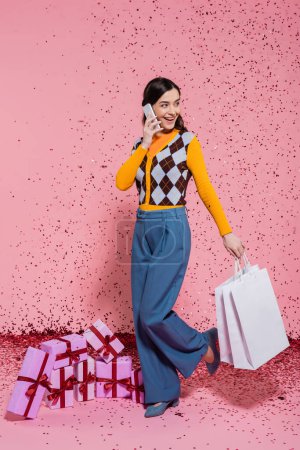 Foto de Mujer sonriente y de moda con bolsas de compras hablando en el teléfono inteligente cerca de cajas de regalo y confeti sobre fondo rosa - Imagen libre de derechos
