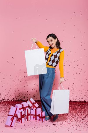longitud completa de la mujer de moda posando con bolsas de compras blancas cerca de cajas de regalo y confeti festivo sobre fondo rosa