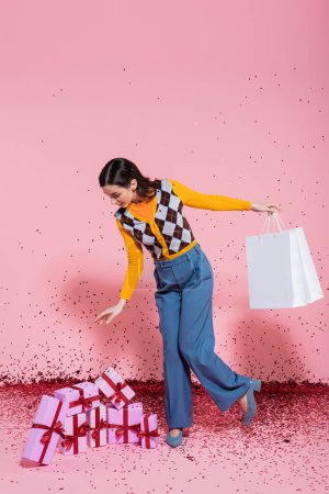 Foto de Longitud completa de la mujer de moda con bolsas de compras mirando cajas de regalo cerca de confeti festivo sobre fondo rosa - Imagen libre de derechos