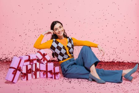 smiling woman in stylish attire sitting near pile of gift boxes and festive confetti on pink background