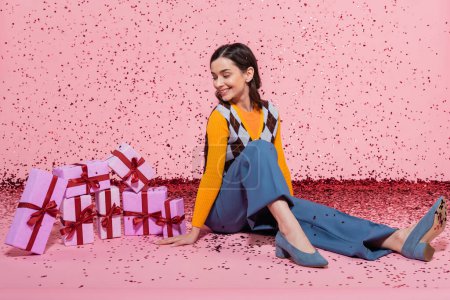 longitud completa de la mujer alegre en ropa de moda mirando la pila de cajas de regalo mientras está sentado cerca de confeti sobre fondo rosa