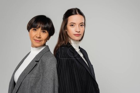 asian mother and daughter in blazers standing back to back and looking at camera isolated on grey