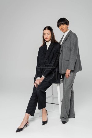 Téléchargez les photos : Full length of middle aged asian woman in stylish suit standing near young daughter sitting on chair on grey background - en image libre de droit