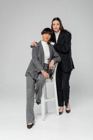 Téléchargez les photos : Full length of young asian woman in black suit embracing stylish mother posing on white chair on grey background - en image libre de droit