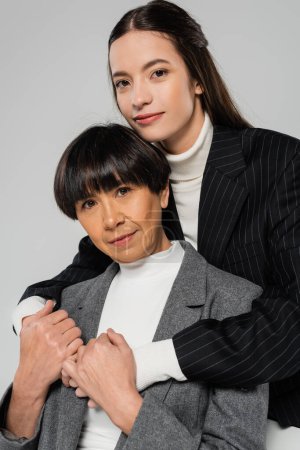 Téléchargez les photos : Brunette asian woman hugging stylish mother and looking at camera isolated on grey - en image libre de droit