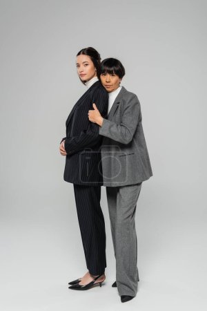 Foto de Full length of happy asian woman in formal wear embracing pretty brunette daughter while looking at camera on grey background - Imagen libre de derechos