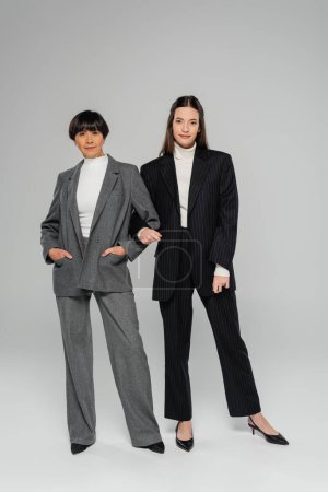 Téléchargez les photos : Full length of positive asian mother and daughter in elegant pantsuits looking at camera on grey background - en image libre de droit