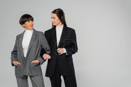 Téléchargez les photos : Asian mother and daughter wearing stylish suits and looking at each other while standing isolated on grey - en image libre de droit