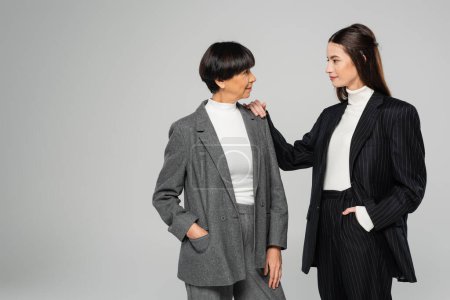 brunette woman in black suit touching shoulder of elegant asian mother isolated on grey