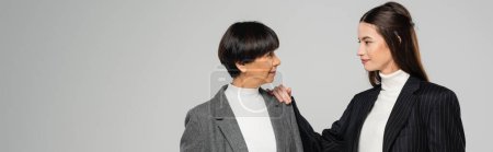 Téléchargez les photos : Positive asian woman in stylish clothes touching shoulder of brunette mother isolated on grey, banner - en image libre de droit