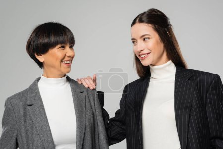 Foto de Cheerful asian mother and daughter in blazers and white turtlenecks looking at each other isolated on grey - Imagen libre de derechos