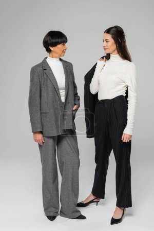 full length of positive asian mother and daughter in formal wear looking at each other while standing on grey background