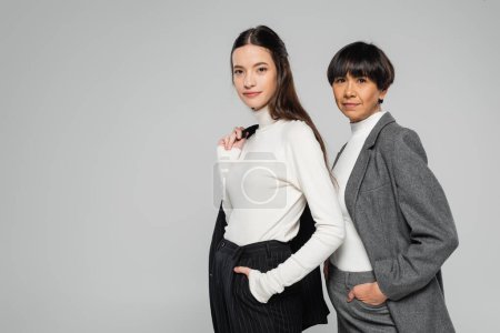 Foto de Positive asian mother and daughter in formal wear posing with hands in pockets while looking at camera isolated on grey - Imagen libre de derechos