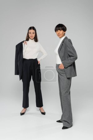 full length of stylish asian mother and daughter in formal wear looking at camera while posing on grey background