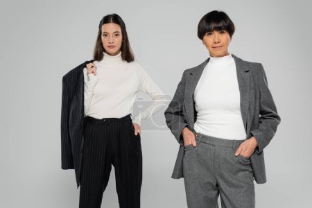 brunette asian mother and daughter in formal wear and white turtlenecks posing and looking at camera isolated on grey