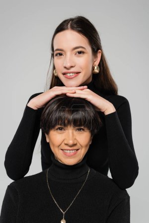 happy and stylish woman in golden earrings posing near middle aged asian mother in black turtleneck isolated on grey