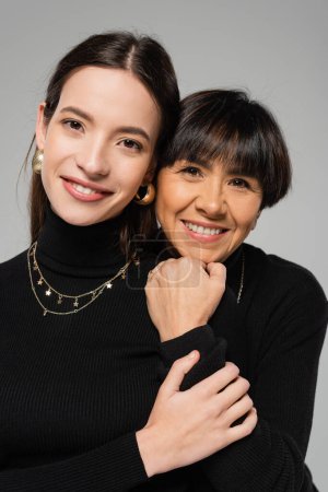portrait of joyful asian mother and daughter in black turtlenecks and necklaces smiling at camera isolated on grey