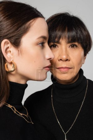 stylish asian woman looking at camera near blurred daughter in black turtleneck and necklaces isolated on grey