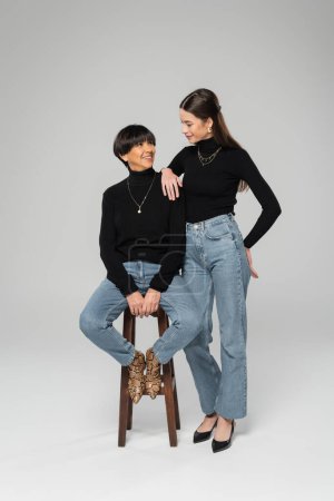 young woman in black turtleneck and jeans smiling at trendy asian mother sitting on wooden stool on grey background