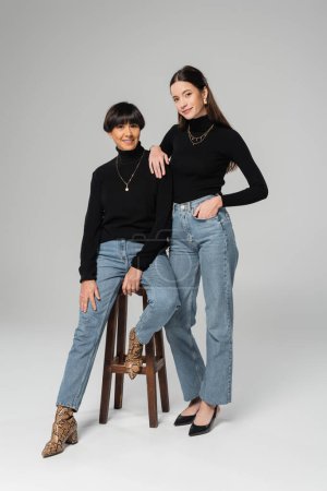 full length of stylish woman standing with hand in pocket of jeans near happy asian mother sitting on wooden stool on grey background