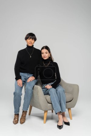 full length of asian mother and daughter in black turtlenecks and jeans posing on armchair on grey background