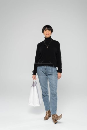 longitud completa de mujer asiática de mediana edad en jeans y botas de piel de serpiente de pie con bolsa de compras sobre fondo gris