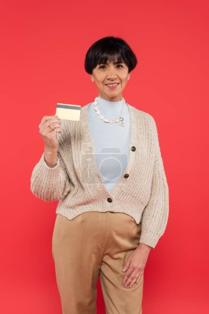 happy asian woman in knitted cardigan and shell beads showing credit card and looking at camera isolated on coral