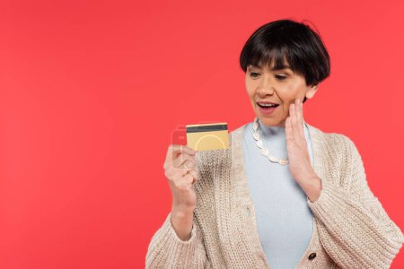amazed asian woman in knitted cardigan and shell beads touching face while looking at credit card isolated on coral