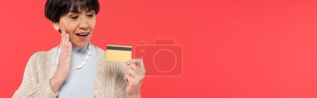 Photo for Impressed asian woman in cardigan touching face while holding credit card isolated on coral, banner - Royalty Free Image