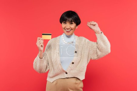 Photo for Brunette asian woman in knitted cardigan holding credit card and celebrating success isolated on coral - Royalty Free Image