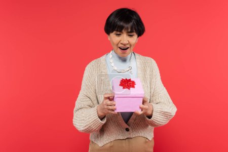 Foto de Surprised asian woman in knitted cardigan looking at gift box with red bow isolated on coral - Imagen libre de derechos